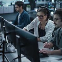 Young Team of Specialists Working on Desktop Computers and Having a Conversation at a Workplace. Female and Male Software Developers Discussing a Solution for Their Artificial Intelligence Project