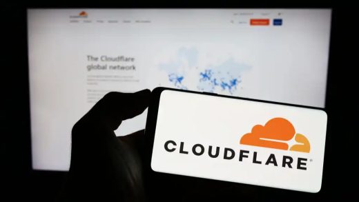 A photograph of a person holding a smartphone with the Cloudflare logo displayed. Behind the phone is a blurred monitor displaying the Cloudflare website.,
