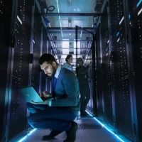 An IT technician works on laptop in data center, with other IT staff in the background.