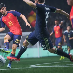 A Spain player in a red shirt strikes the ball toward goal as a defender in navy lunges to block