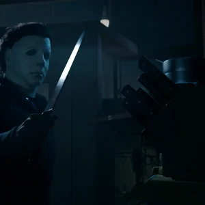 Michael Myers standing in a dark kitchen inspecting a long knife taken from a wooden knife block