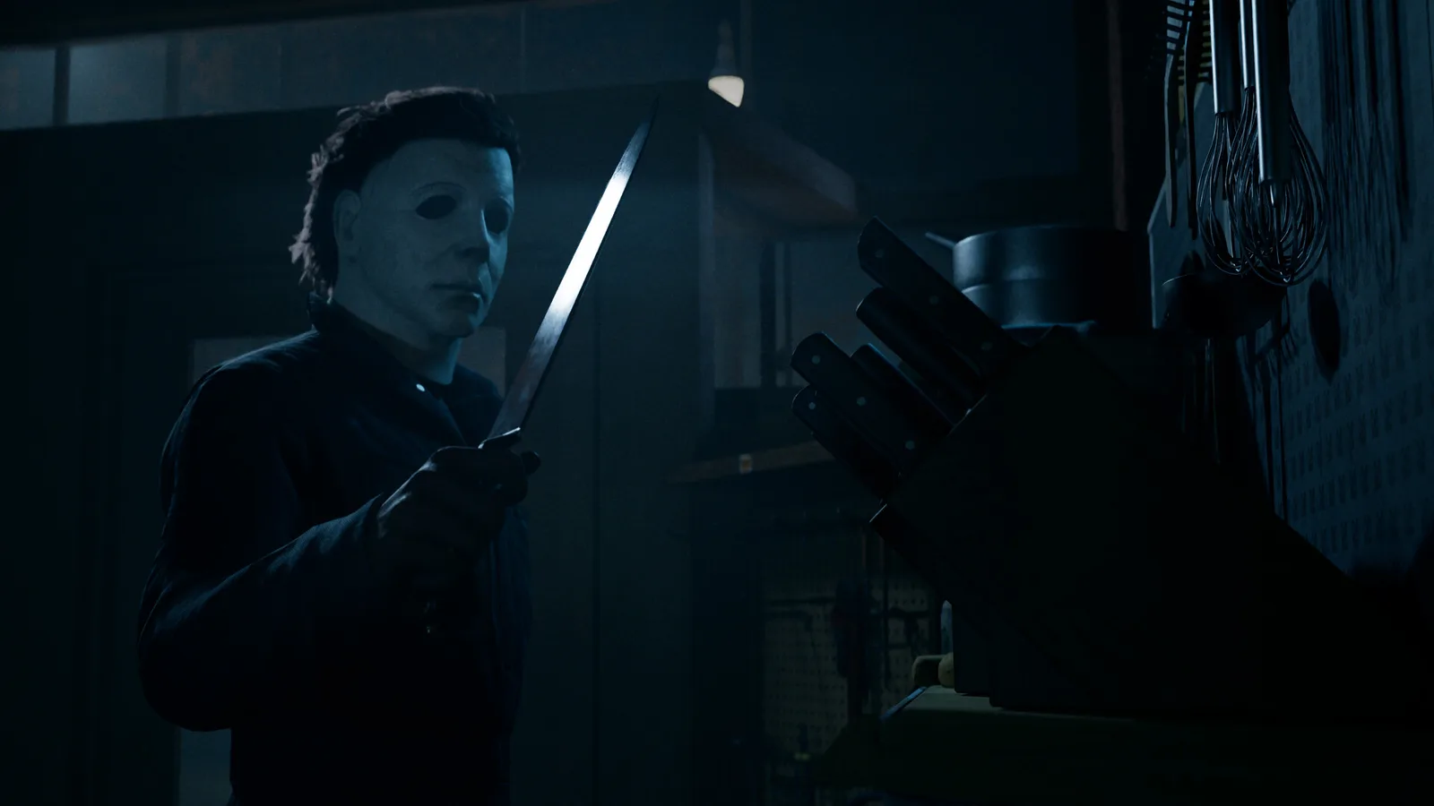 Michael Myers standing in a dark kitchen inspecting a long knife taken from a wooden knife block