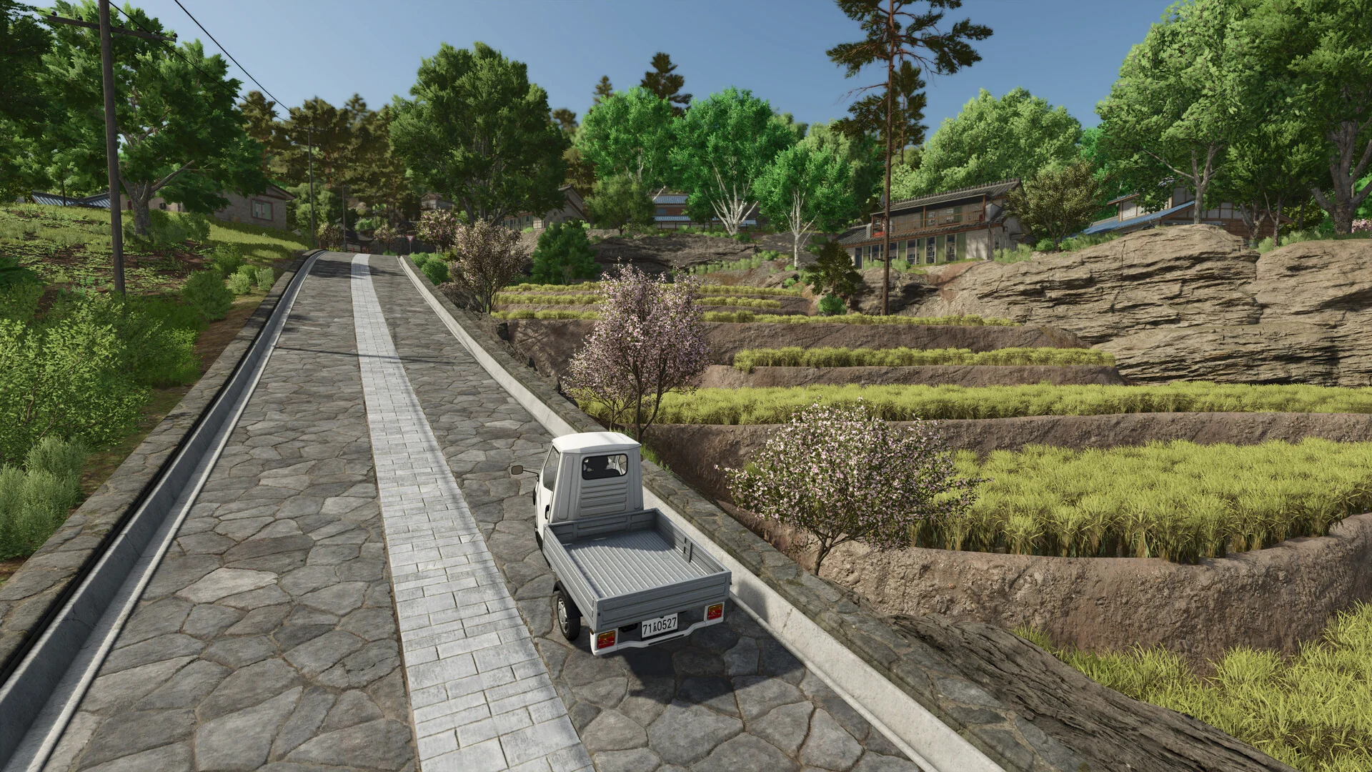 A small white pickup truck drives up a stone road past terraced fields and village houses in Farming Simulator 25