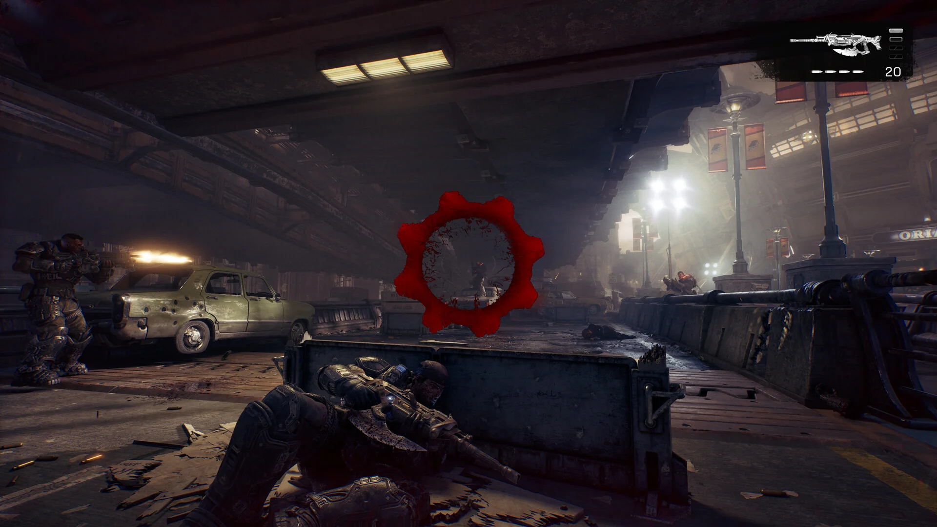 Third-person gameplay with a soldier in cover behind a barrier aiming at enemies under an elevated rail line, with the red damage indicator in the middle of the screen