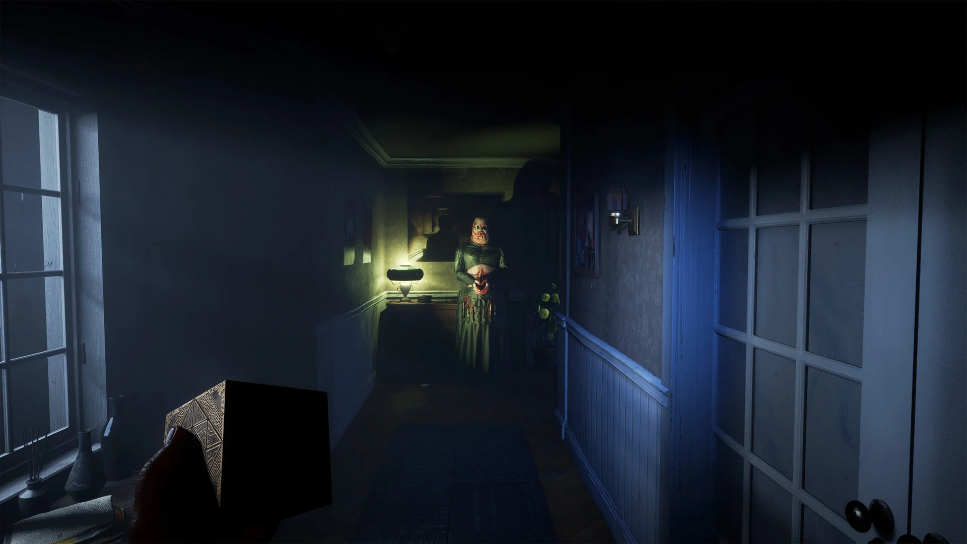 A dark house hallway lit by one wall lamp, with a figure standing at the far end beside a doorway