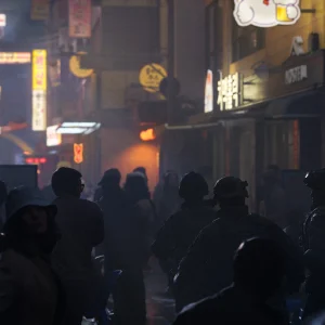 A crowd of people in silhouette fills a narrow alley lit by neon shop signs at night