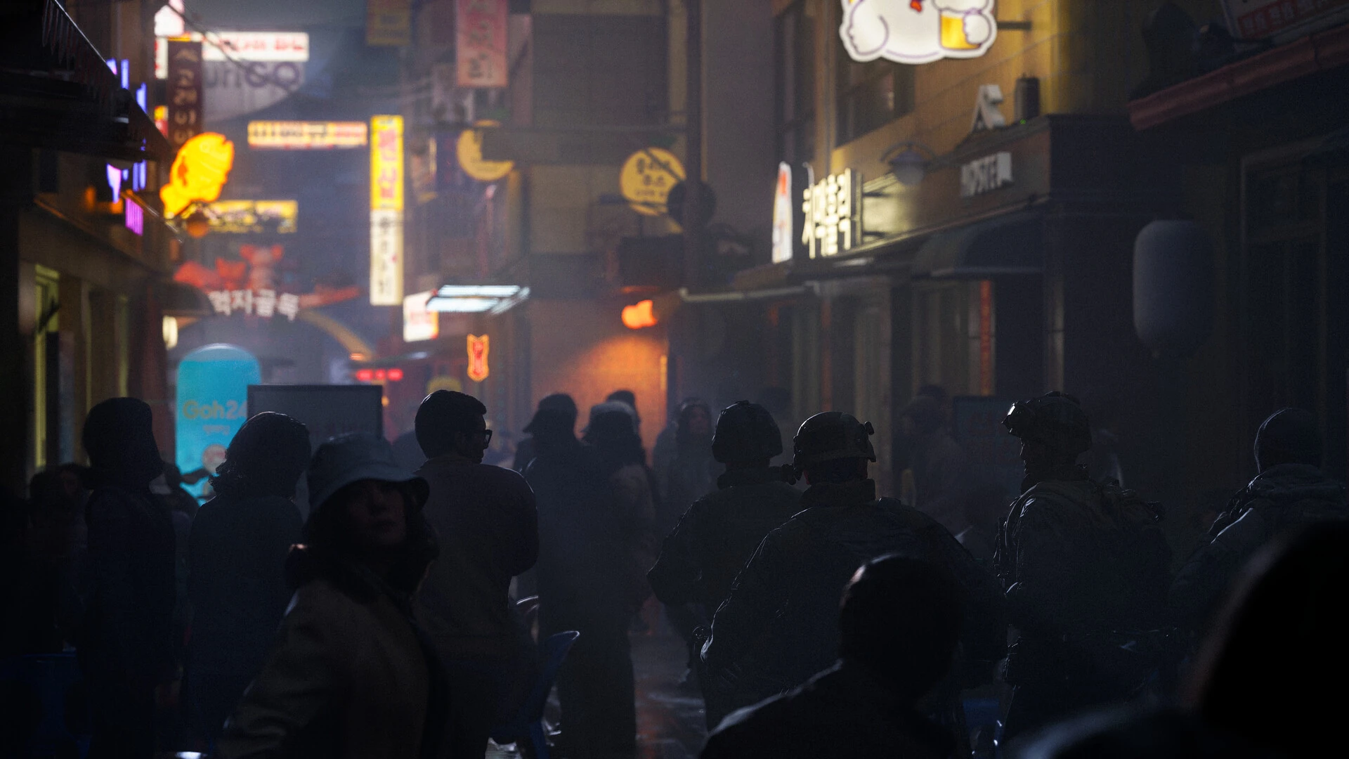 A crowd of people in silhouette fills a narrow alley lit by neon shop signs at night
