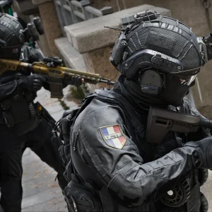 Two operators in black tactical helmets with French flag patches aim rifles beside an armored vehicle on a city street
