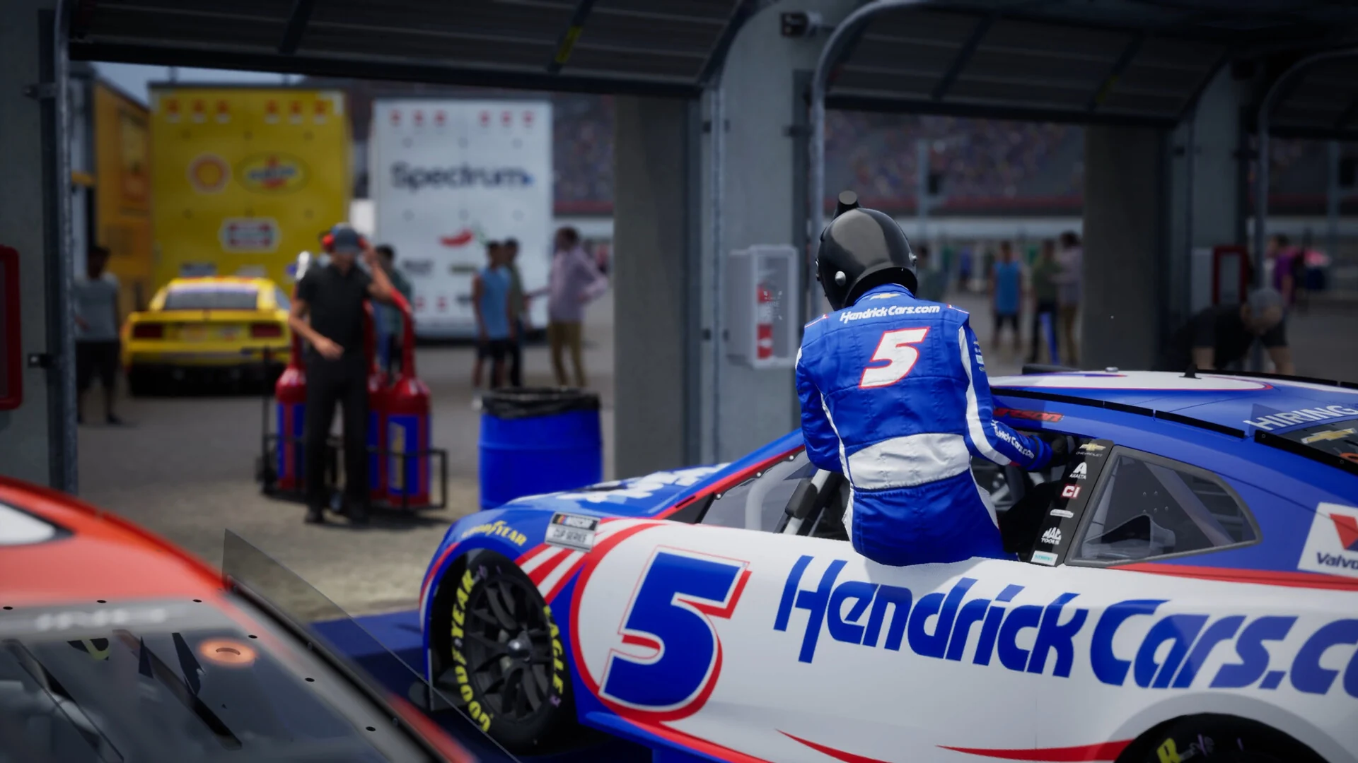 A driver in a blue firesuit climbs through the window of the No. 5 Hendrick Cars Chevrolet in a garage stall while crew members work behind it