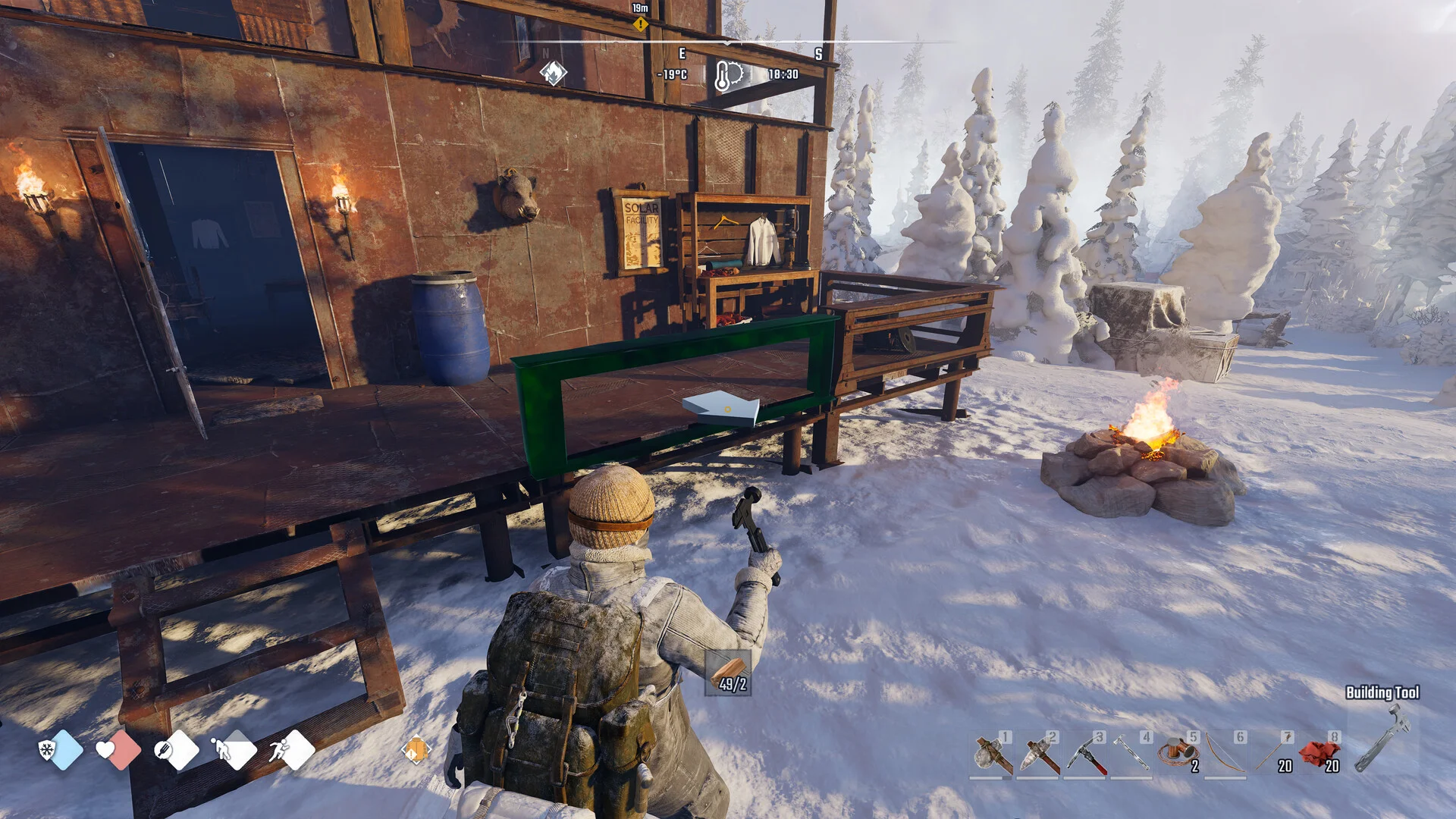 Third-person view of a survivor placing wooden walls for a cabin next to a campfire in the snow, with the building hotbar at the bottom of the screen