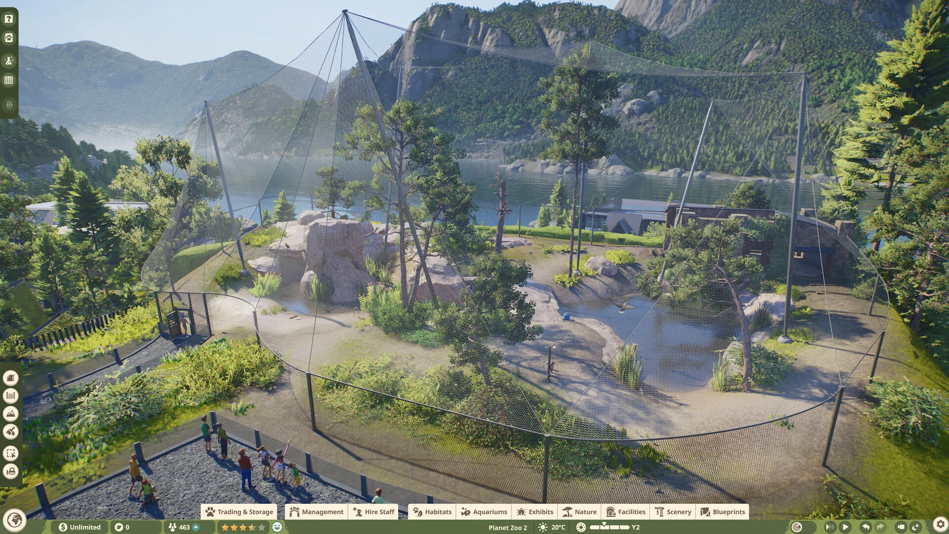 A large walk-through aviary in Planet Zoo 2 with a tall mesh cover over trees, rocks and a pond, seen from above