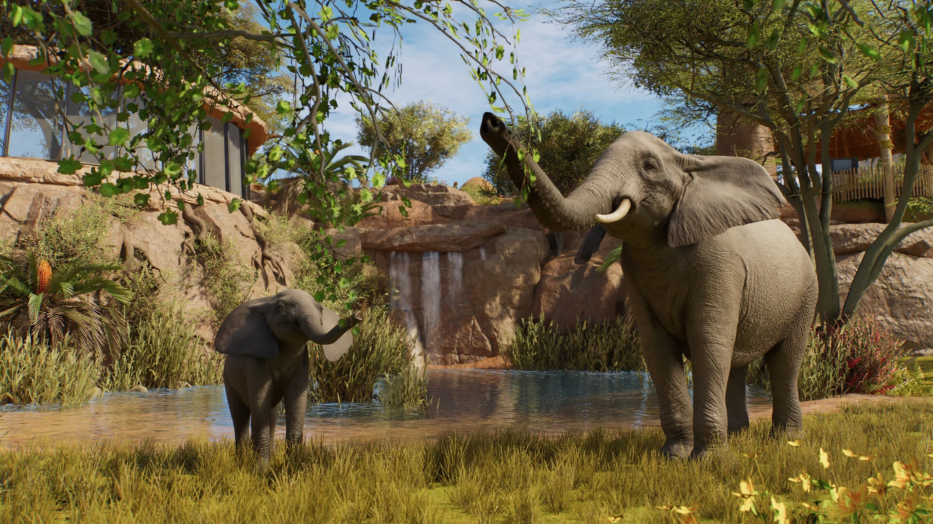 An adult elephant raises its trunk beside a calf in a grassy Planet Zoo 2 habitat with rocks and a small waterfall