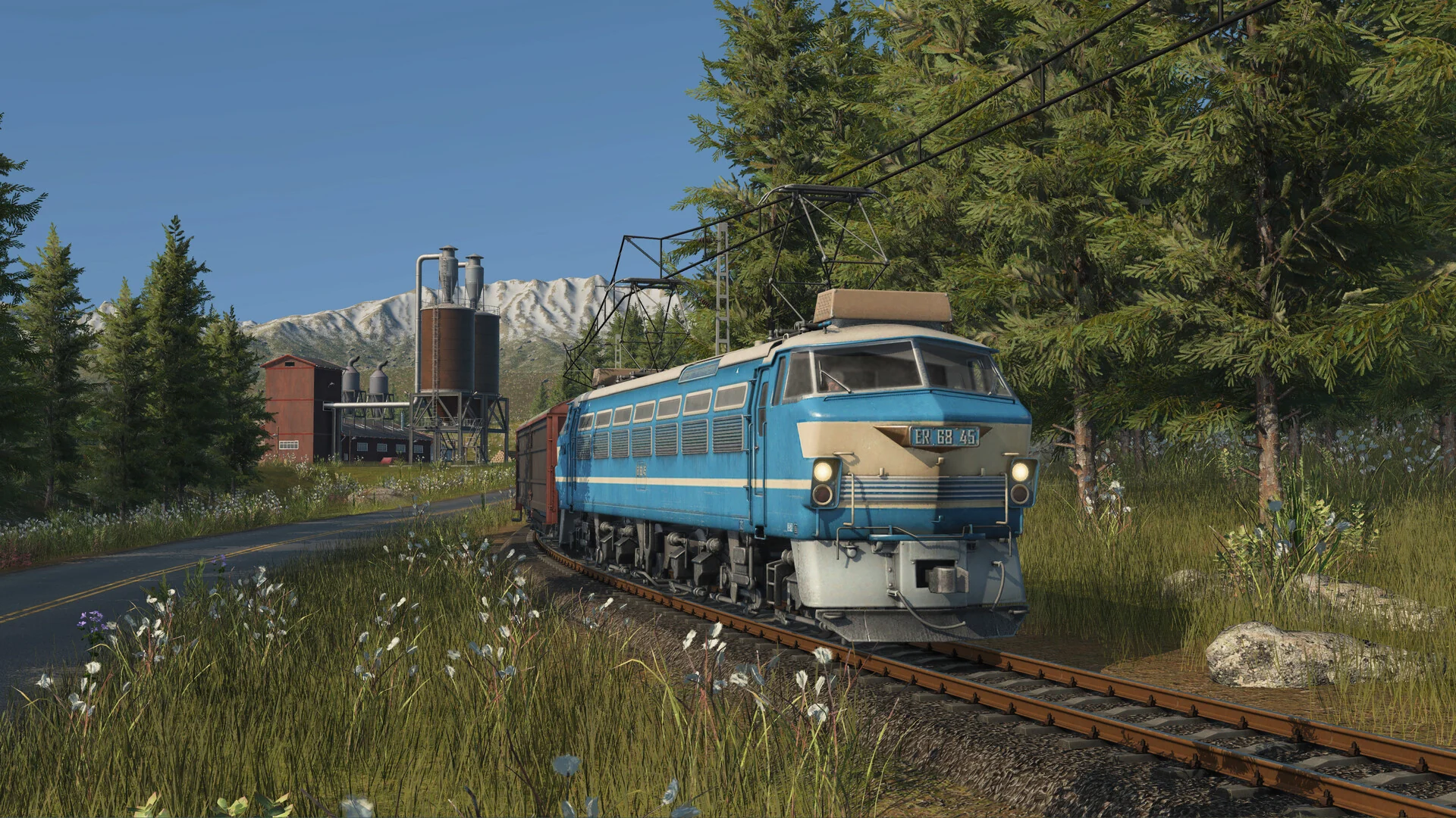 A blue and cream electric locomotive running under overhead wires through a pine forest, with snowy mountains behind it in Transport Fever 3