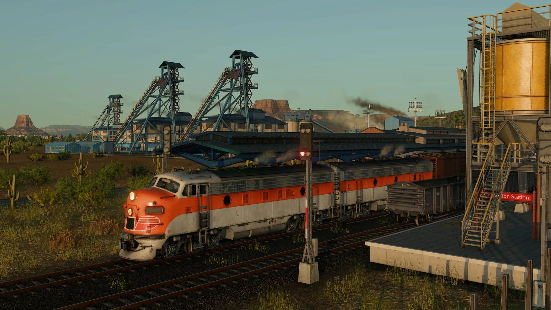 A vintage orange and silver diesel locomotive pulling freight wagons past coal mine headframes and a desert station in Transport Fever 3